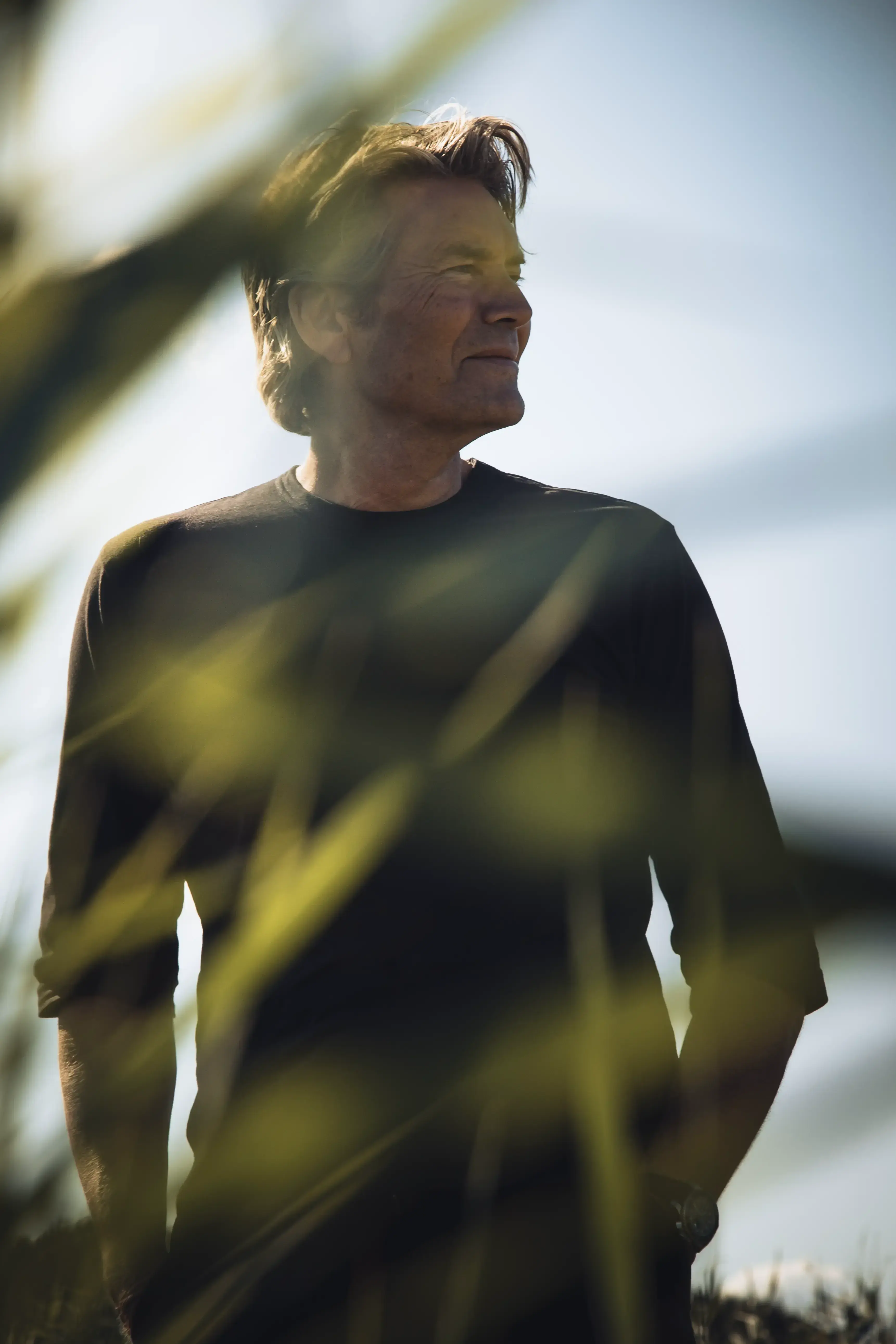 Piet Boon in a black shirt stands outdoors, looking to the side, with blurred green foliage in the foreground and a clear blue sky.